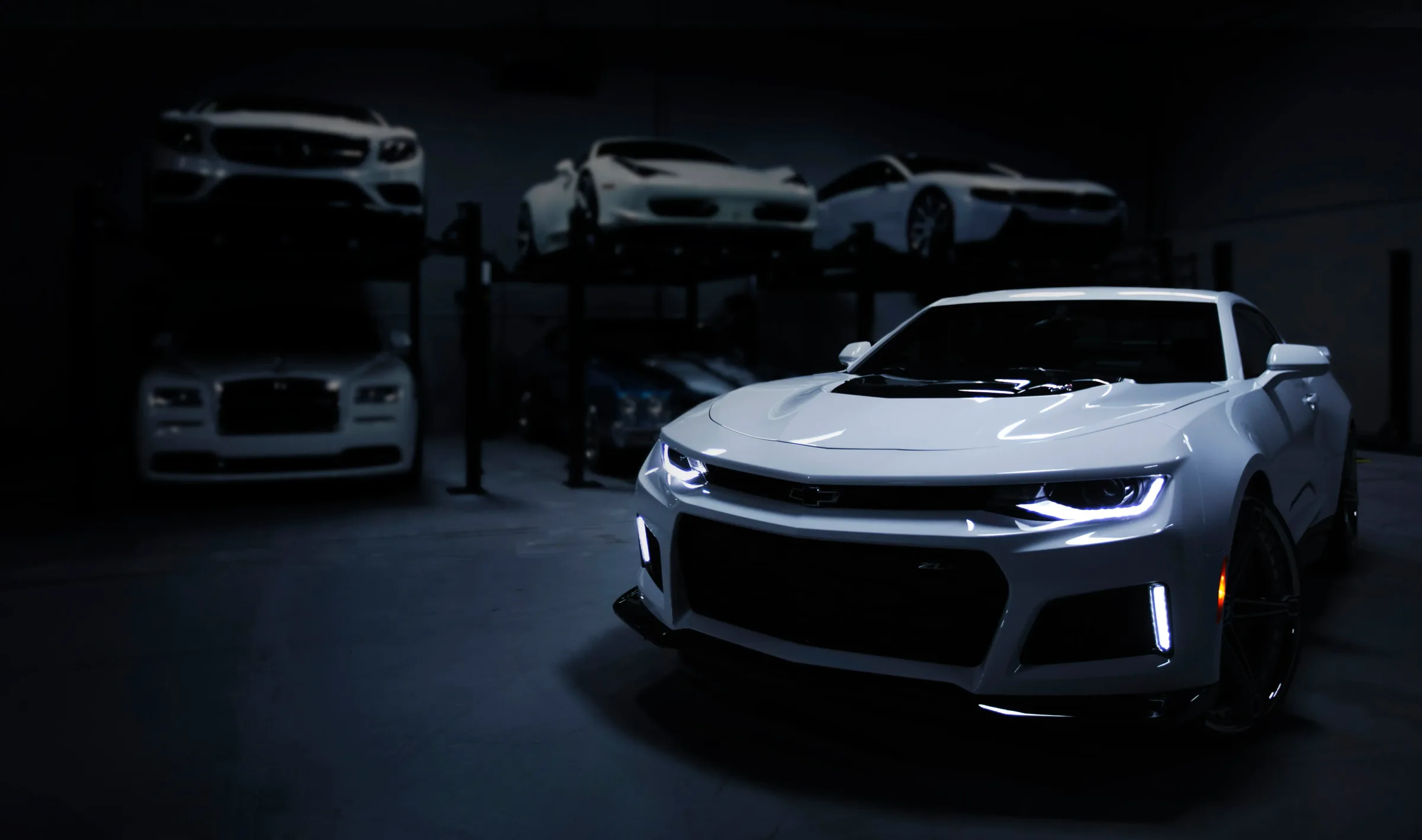 A white performance car lit in a dark workshop, with further vehicles raised on lifts behind it